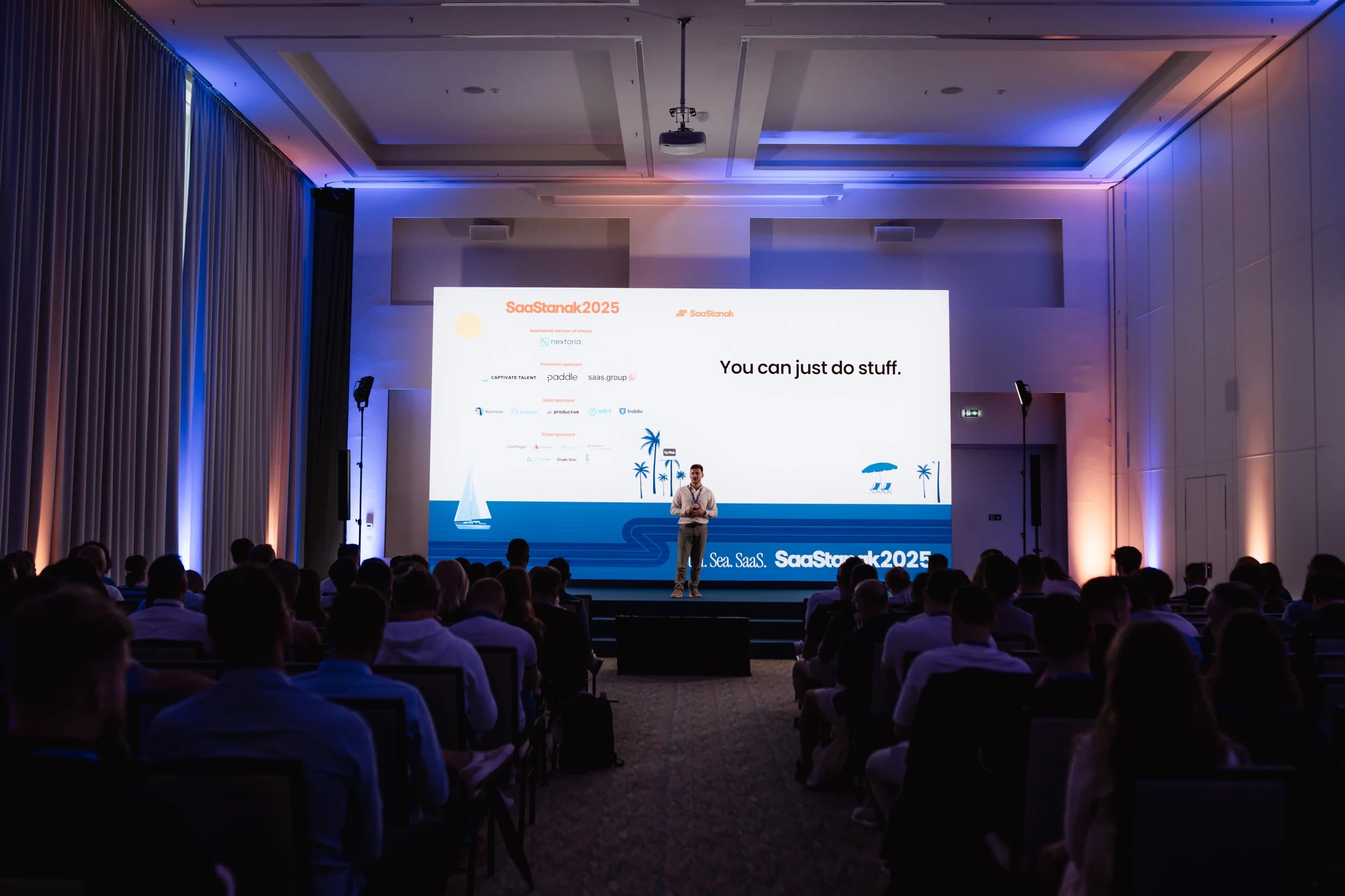 A man speaking on a stage at SaaStanak 2025 with a large screen behind him saying 'You can just do stuff'